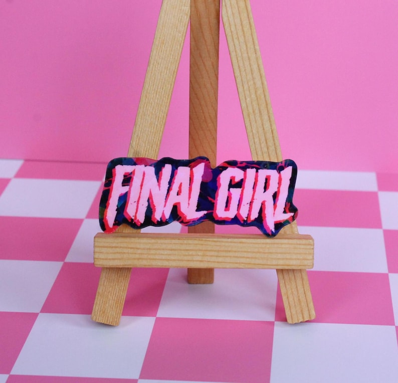 Pu&ograve; includere: Un adesivo rosa e blu "Final Girl" esposto su un piccolo cavalletto di legno. L'adesivo presenta le parole "FINAL GIRL" in rosa, delineate in un colore pi&ugrave; scuro, su uno sfondo blu e viola. Lo sfondo &egrave; un motivo a scacchi rosa e bianchi.