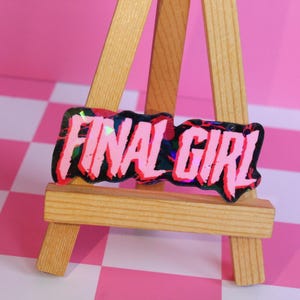 Pu&ograve; includere: Un adesivo rosa e nero con le parole "FINAL GIRL" in un carattere audace e stilizzato. L'adesivo ha un effetto olografico iridescente ed &egrave; esposto su un piccolo cavalletto di legno su uno sfondo a scacchi rosa e bianco.