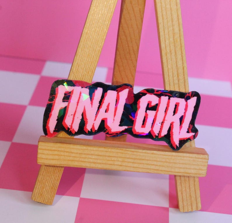 Pu&ograve; includere: Un adesivo rosa e nero con le parole "FINAL GIRL" in un carattere audace e stilizzato. L'adesivo ha un effetto olografico iridescente ed &egrave; esposto su un piccolo cavalletto di legno su uno sfondo a scacchi rosa e bianco.