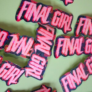 Pu&ograve; includere: Adesivi rosa e neri glitterati con le parole "FINAL GIRL" in un carattere audace e stilizzato. Gli adesivi hanno un'estetica usurata e spigolosa, sparsi su una superficie verde chiaro.