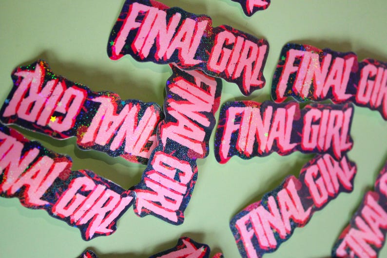 Pu&ograve; includere: Adesivi rosa e neri glitterati con le parole "FINAL GIRL" in un carattere audace e stilizzato. Gli adesivi hanno un'estetica usurata e spigolosa, sparsi su una superficie verde chiaro.