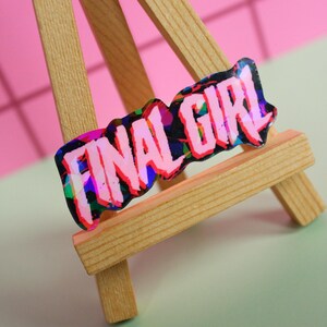Pu&ograve; includere: Un adesivo olografico con la scritta "FINAL GIRL" in rosa, nero e blu, esposto su un piccolo cavalletto di legno. Lo sfondo &egrave; un mix di rosa e verde.