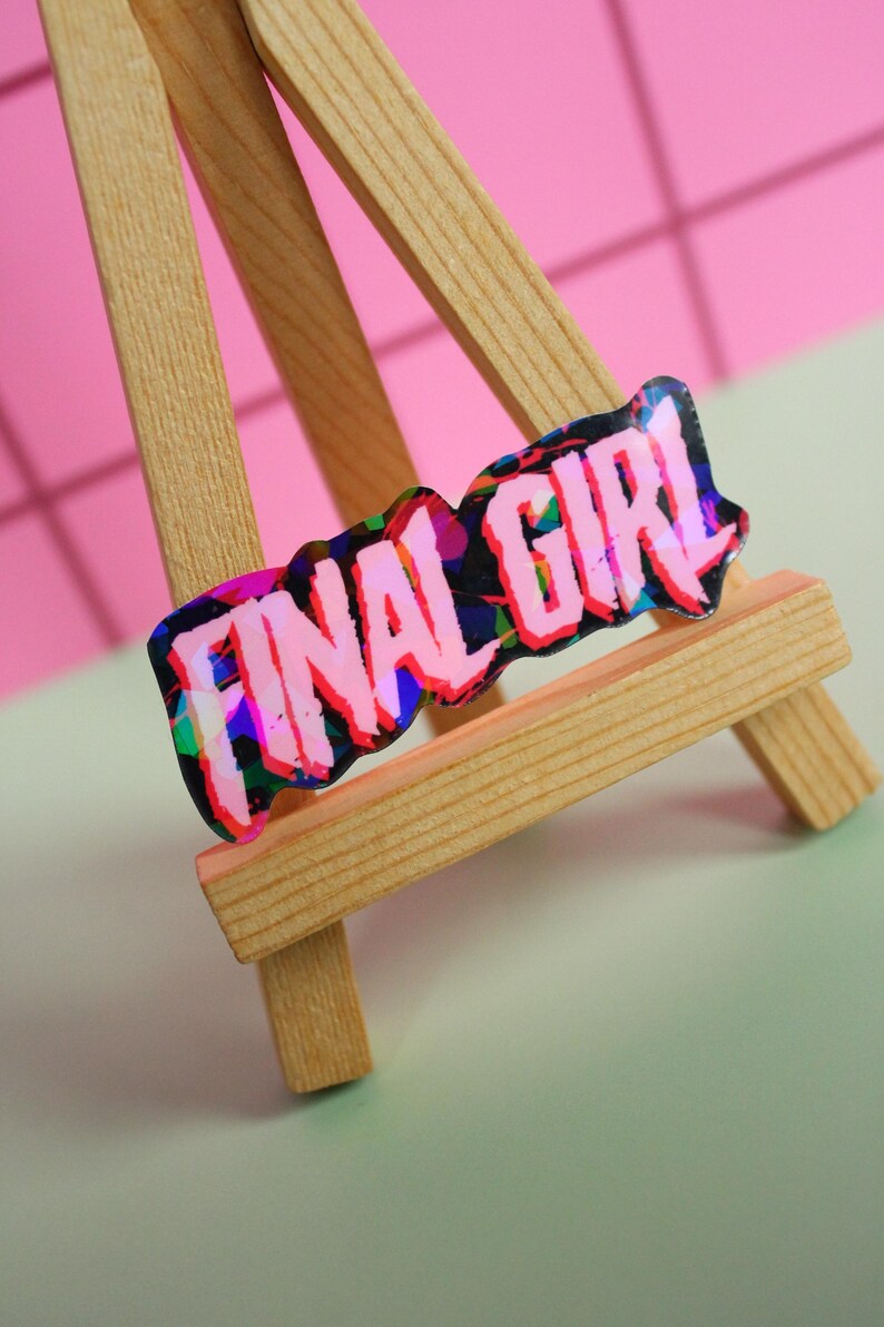 Pu&ograve; includere: Un adesivo olografico con la scritta "FINAL GIRL" in rosa, nero e blu, esposto su un piccolo cavalletto di legno. Lo sfondo &egrave; un mix di rosa e verde.
