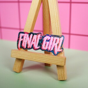 Pu&ograve; includere: Un cavalletto in miniatura in legno espone un adesivo rettangolare con la scritta "FINAL GIRL" in rosa, delineata in un colore pi&ugrave; scuro. L'adesivo ha un effetto glitterato. Lo sfondo &egrave; una parete rosa e una superficie verde.
