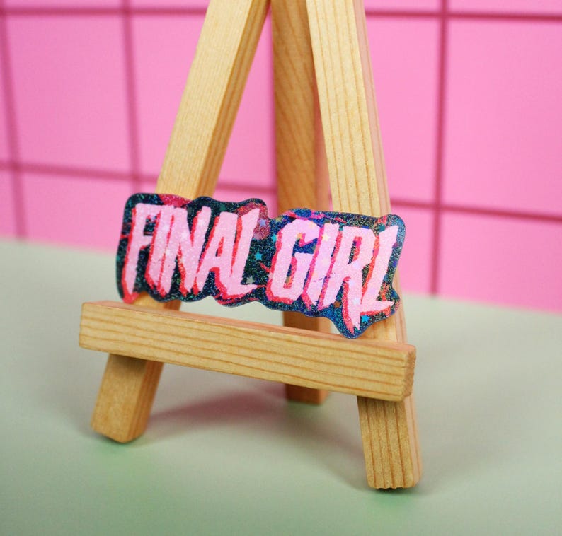 Pu&ograve; includere: Un cavalletto in miniatura in legno espone un adesivo rettangolare con la scritta "FINAL GIRL" in rosa, delineata in un colore pi&ugrave; scuro. L'adesivo ha un effetto glitterato. Lo sfondo &egrave; una parete rosa e una superficie verde.