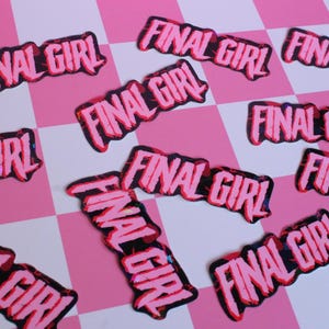 Pu&ograve; includere: Una collezione di toppe rosa e nere "FINAL GIRL" sparse su una superficie a scacchi rosa e bianca. Le toppe hanno un carattere audace e stilizzato con un contorno nero e un riempimento rosa.