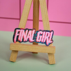 Pu&ograve; includere: Un piccolo cavalletto di legno espone una toppa rettangolare con le parole "FINAL GIRL" in rosa, bordate di blu e nero. Lo sfondo &egrave; un muro rosa piastrellato e una superficie verde chiaro.