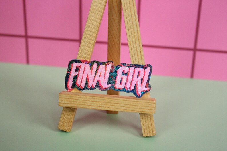 Pu&ograve; includere: Un piccolo cavalletto di legno espone una toppa rettangolare con le parole "FINAL GIRL" in rosa, bordate di blu e nero. Lo sfondo &egrave; un muro rosa piastrellato e una superficie verde chiaro.