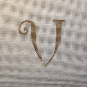 May include: Close-up of a canvas with a gold-colored letter "V" embroidered on it. The letter has a decorative, cursive style with a curled top and a pointed bottom. The canvas has a fine, woven texture and a neutral, off-white color.