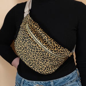 May include: A leopard print fanny pack with a gold zipper and a cream-colored strap. The bag is worn across the body. The bag's pattern is a mix of black spots on a tan background.