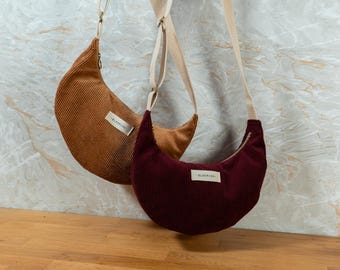 May include: Two crescent-shaped corduroy bags in shades of brown and burgundy hang against a marble-patterned wall. Each bag has a matching strap and a small label that reads "BLOOMING". The bags are a stylish accessory.