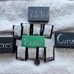 May include: A collection of small, dark wooden boxes and packets arranged on a white quilted surface. The boxes are labeled "Wicked", "Hexes", and "Curses" in silver lettering. A green sign reads "Poison".