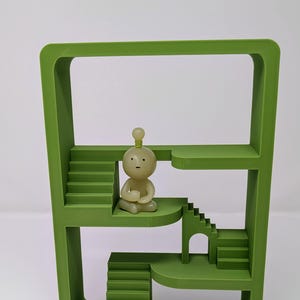 May include: A green, geometric sculpture featuring multiple levels with staircases and a small figure in a meditative pose. The sculpture has a modern design and is set against a white background.