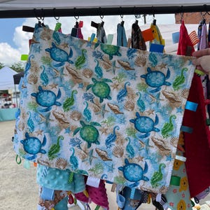 May include: A baby blanket with a sea turtle pattern in shades of blue and green, featuring seashells and starfish on a white background. The blanket is rectangular and has colourful ribbons attached to the edges. Other blankets are visible in the background.