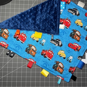 Puede incluir: Una manta para bebé azul con un estampado de coches de dibujos animados y una tela azul marino con hoyuelos en un lado. La manta tiene cintas de colores unidas a los bordes.