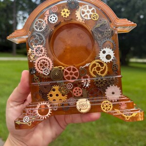 May include: A translucent amber-colored resin clock frame featuring an embedded assortment of metallic gears, cogs, and clock hands in various sizes, shapes, and finishes, including gold, silver, copper, and bronze tones, arranged around a central circular void.
