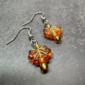 May include: A pair of dangle earrings featuring amber-colored, leaf-shaped beads. The earrings have silver-colored ear wires and small, round beads. The leaf beads have intricate vein detailing, and the overall design is autumnal.