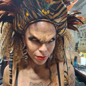 Puede incluir: Un tocado decorativo con un estampado de rayas de tigre marrón y negro, plumas y detalles dorados. La persona tiene ojos naranjas, lápiz labial rojo y pendientes de serpiente. Un tatuaje de mariposa es visible en el pecho.