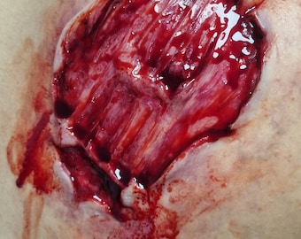 May include: A gruesome special effects makeup piece depicting a deep, open wound on a shoulder. The wound is filled with realistic-looking blood and exposed tissue, creating a disturbing visual effect. The skin around the wound is pale.