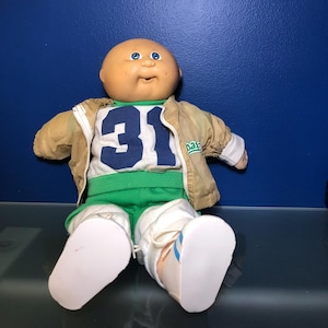 May include: A vintage Cabbage Patch Kid doll wearing a tan jacket, a white and blue jersey with the number 31, green shorts, and white shoes. The doll has a bald head, blue eyes, and a smiling expression.