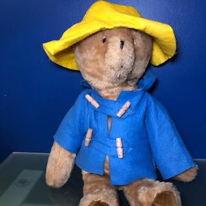 May include: A plush teddy bear wearing a bright yellow hat and a blue coat with wooden toggle closures. The bear has brown fur and is sitting on a reflective surface against a blue background.