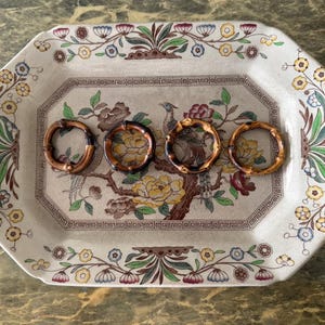 Può includere: Un vassoio da portata ottagonale in ceramica con un motivo floreale e di uccelli, con un'immagine centrale di un uccello e anelli portatovaglioli in bambù. Il vassoio ha uno sfondo color crema con bordi floreali colorati.