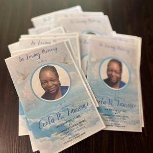 May include: A stack of funeral program booklets featuring a light blue sky background with white clouds and doves. Each booklet displays the text In Loving Memory, the name Ceola M. Lawrence, dates, and service details alongside a circular portrait photo.