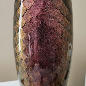 May include: A decorative, oval-shaped tumbler with a textured, scale-like pattern. The color gradient transitions from gold to burgundy, creating a shimmering effect. The tumbler is likely used for beverages.