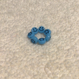 May include: A close-up of a blue smiley face bead bracelet. The bracelet is made of small, round beads with a smiley face design. The beads are strung together on a clear elastic cord.