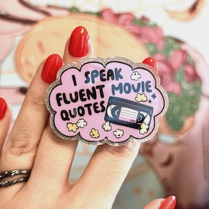 May include: A pink, cloud-shaped pin with the text "I SPEAK FLUENT MOVIE QUOTES" and a VHS tape illustration. The pin has a glittery silver border and small popcorn and cloud graphics. The background is a soft, blurred pattern.