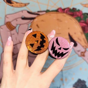 May include: Two Halloween-themed phone grips, one orange with glitter and one pink with glitter, both featuring a jack-o'-lantern face.