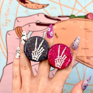 May include: Two round phone grips with glittery surfaces. One is black with a white skeleton hand in a peace sign, the other is pink with a white skeleton hand. The background is a light blue fabric with the word "Sagittarius".
