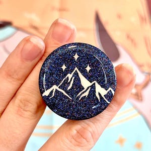 May include: A round, dark blue glittery phone grip featuring a white mountain range and three white stars. The phone accessory is held in a hand, showcasing the design's details.