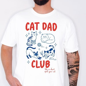 May include: White t-shirt featuring a red and blue graphic with the text "CAT DAD CLUB" and cat illustrations. The shirt also has the text "life is short, spoil your cat". The t-shirt is made of a soft material.