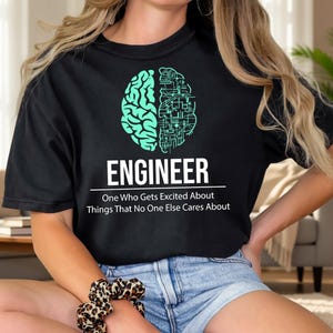 May include: Black t-shirt with a teal brain graphic and the word "ENGINEER" in white. The text below reads "One Who Gets Excited About Things That No One Else Cares About". The shirt is a casual style.