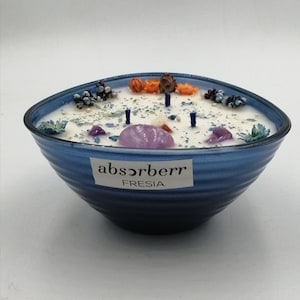 Pode incluir: Uma vela de vidro azul em forma de tigela com uma base de cera branca. A vela é decorada com pequenas flores, cristais e um rótulo que diz "absorberr FRESIA".
