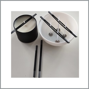 May include: A black candle jar with a white candle inside and a black wick. A white bowl with four black wicks and metal bases. Four additional black wicks are on a white surface.