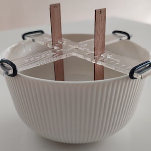 May include: A white ceramic bowl with a clear plastic measuring tool with wooden dowels. The tool has a ruler on each side and the dowels are labeled "12".