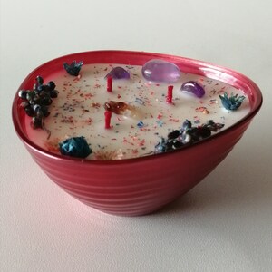 Pode incluir: Uma vela em forma de tigela vermelha com uma base de cera branca e cristais, flores secas e glitter incrustados. A vela tem vários pavios.