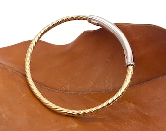 May include: A gold and silver bangle bracelet with a twisted design. The bracelet is on a brown leaf.