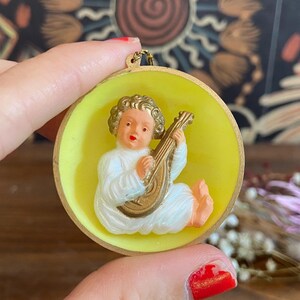 May include: A vintage-style pendant featuring a cherubic figure playing a lute. The figure is in white robes, set against a yellow background within a gold-toned frame. The pendant is held by a hand, with a colorful background.