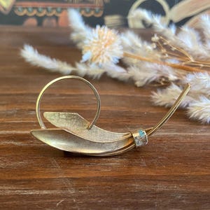 May include: A gold-toned brooch with a unique, abstract design. The brooch features layered, leaf-like shapes and a circular element. A small, turquoise-colored accent is set near the pin's end. The brooch is displayed on a wooden surface.