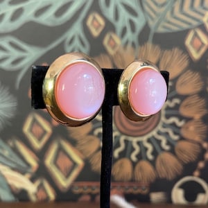 May include: A pair of vintage clip-on earrings. Each earring features a large, round, light pink cabochon stone set in a gold-tone metal frame. The earrings are displayed on a black stand against a patterned background.