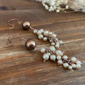 Puede incluir: Un par de pendientes colgantes de color cobre. Cada pendiente presenta una gran cuenta redonda de color bronce en la parte superior, con una cascada de pequeñas perlas de imitación blanquecinas colgando debajo. Los pendientes descansan sobre una superficie de madera.