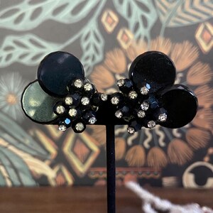 May include: Black vintage clip-on earrings with a cluster of small, sparkling rhinestones and black circular accents. The earrings are displayed on a black stand against a patterned background.