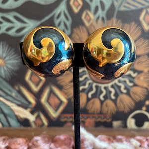 Puede incluir: Un par de pendientes vintage de clip. Cada pendiente es una esfera azul marino oscuro con un diseño dorado en espiral. Los pendientes se exhiben en un soporte negro sobre un fondo estampado.