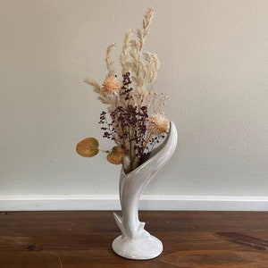 May include: A white ceramic vase with a unique curved design, holding a bouquet of dried flowers with brown, white, and purple accents.
