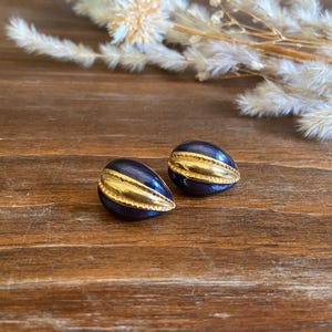 May include: A pair of vintage clip-on earrings. Each earring has a dark blue, oval base with a gold-coloured, horizontal band across the centre. The earrings are on a wooden surface with dried floral accents.