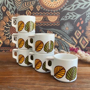 Może przedstawiać: Sześć białych ceramicznych kubków ułożonych w stos, każdy z wzorem liści w kolorze żółtym, zielonym i brązowym. Kubki mają uchwyt i stoją na drewnianej powierzchni. Tło przedstawia wzór kwiatowy.