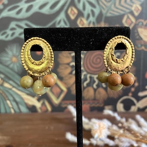 Könnte beinhalten: Ein Paar goldfarbene Ohrringe mit ovalem Design und einer Anhänger-Anordnung aus kleinen, runden Perlen in Grün- und Brauntönen. Die Ohrringe werden auf einem schwarzen Ständer vor einem gemusterten Hintergrund präsentiert.